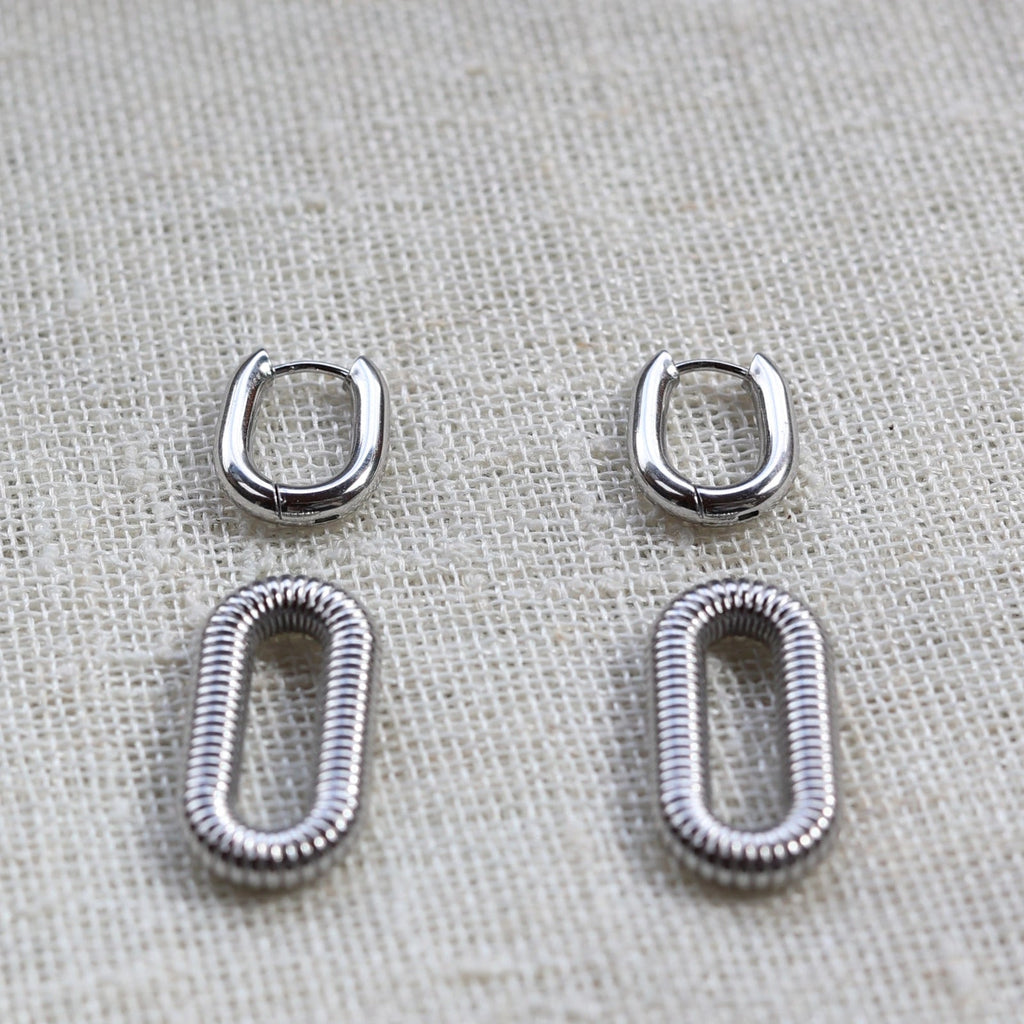 Boucles d'oreilles "J'existe" en acier argenté, au design modulable, photographiées sur un tissu beige texturé.