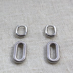 Boucles d'oreilles "J'existe" en acier argenté, au design modulable, photographiées sur un tissu beige texturé.
