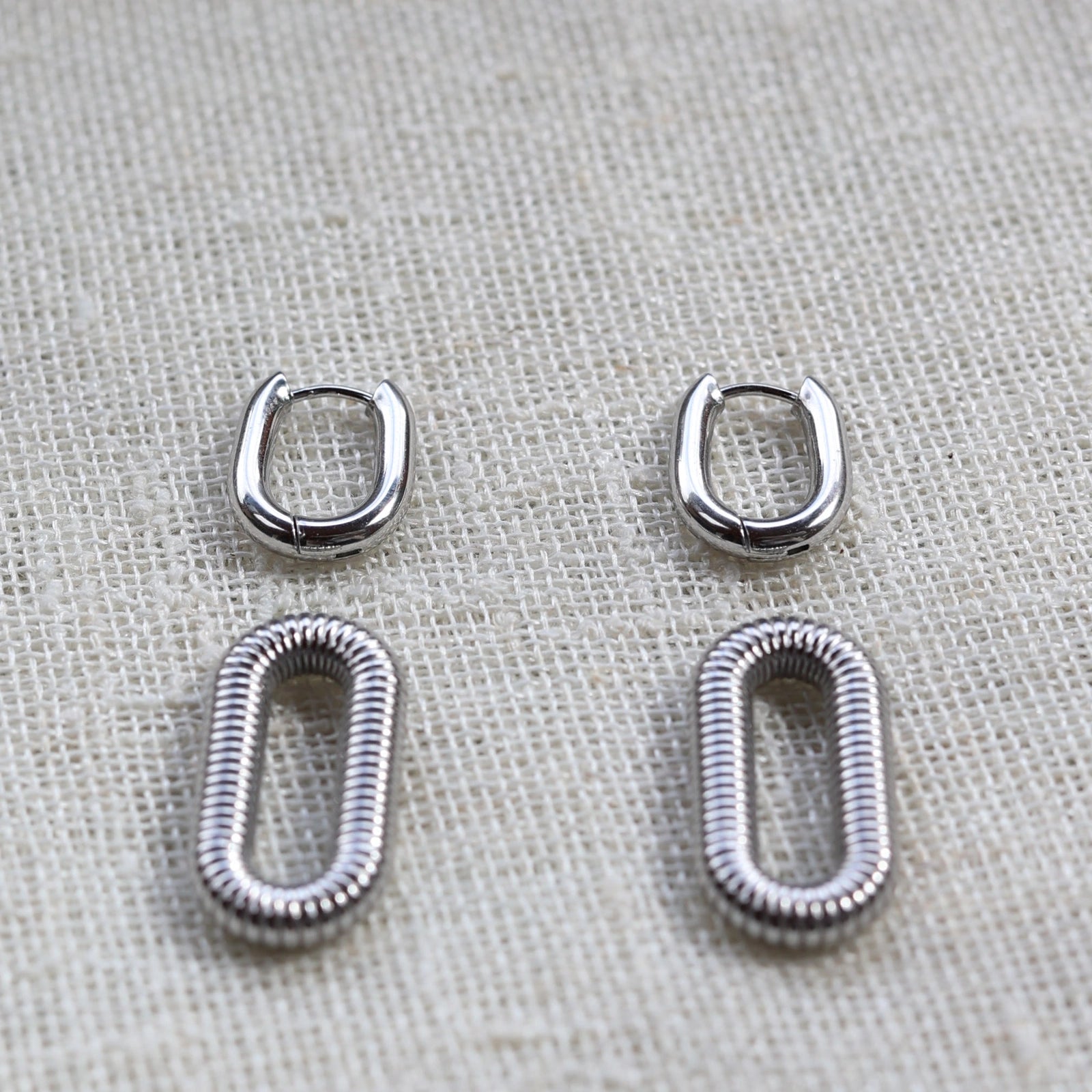 Boucles d'oreilles "J'existe" en acier argenté, au design modulable, photographiées sur un tissu beige texturé.