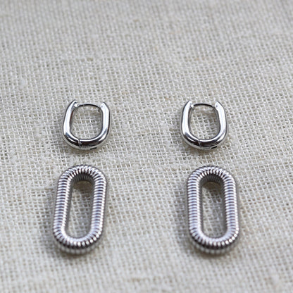 Boucles d'oreilles "J'existe" en acier argenté, au design modulable, photographiées sur un tissu beige texturé.