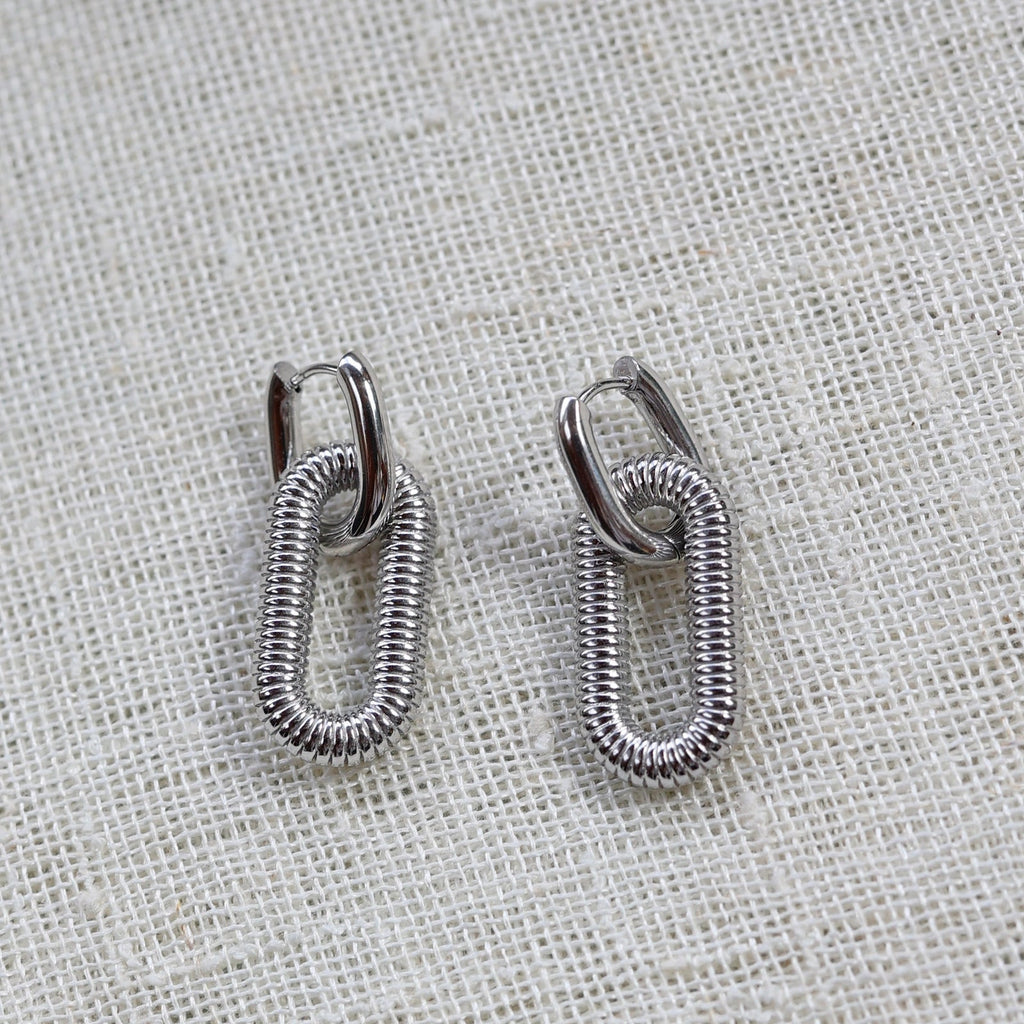 Boucles d'oreilles "J'existe" en acier argenté, au design modulable, photographiées sur un tissu beige texturé.