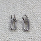 Boucles d'oreilles "J'existe" en acier argenté, au design modulable, photographiées sur un tissu beige texturé.