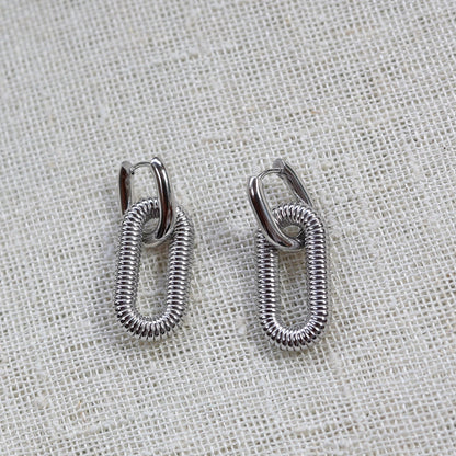 Boucles d'oreilles "J'existe" en acier argenté, au design modulable, photographiées sur un tissu beige texturé.