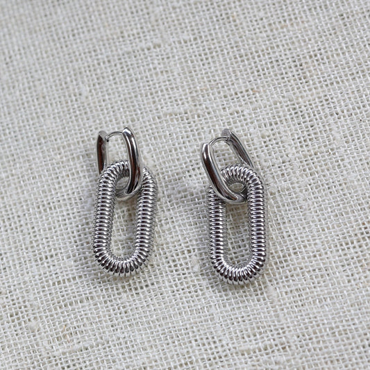 Boucles d'oreilles "J'existe" en acier argenté, au design modulable, photographiées sur un tissu beige texturé.
