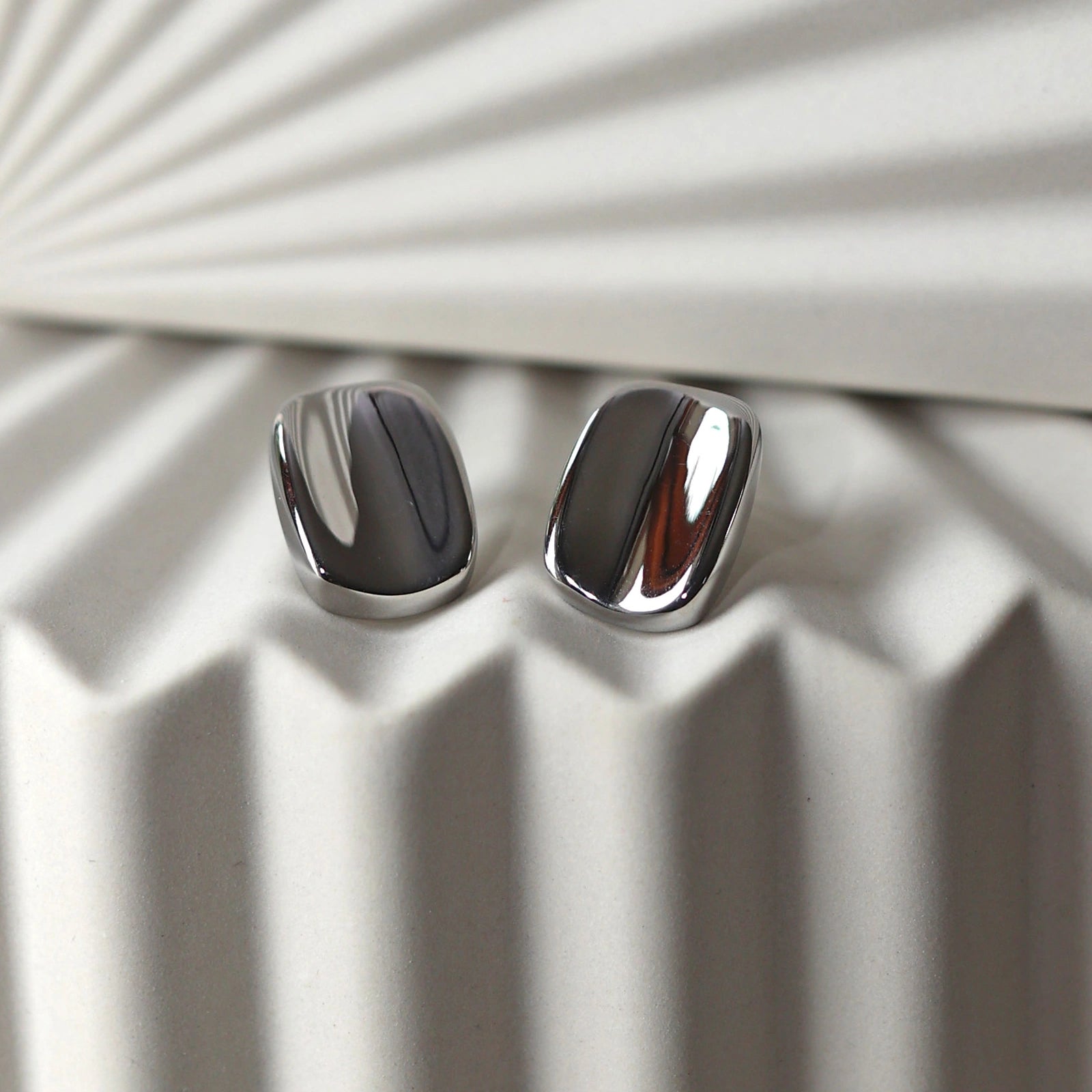 Zoom sur les boucles d’oreilles "Fierté" minimalistes rectangulaires en acier inoxydable argenté, posées à plat sur un support beige texturé.