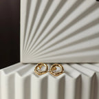 Zoom sur les boucles d'oreilles martelées Source en acier inoxydable doré, posées sur un fond texturé beige.