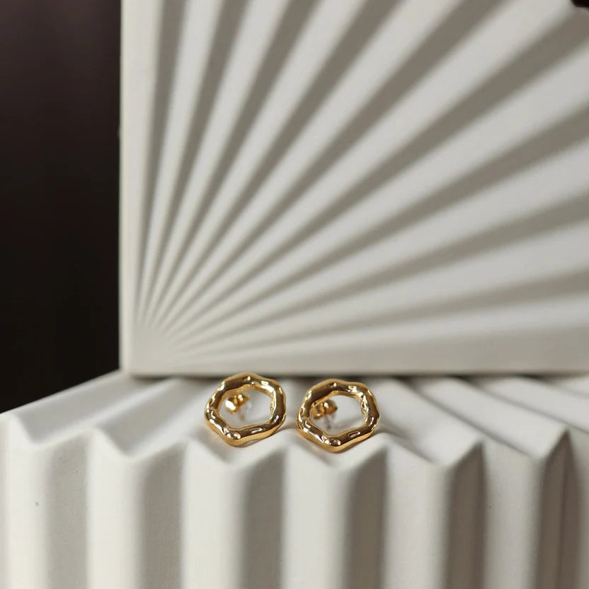 Zoom sur les boucles d'oreilles martelées Source en acier inoxydable doré, posées sur un fond texturé beige.