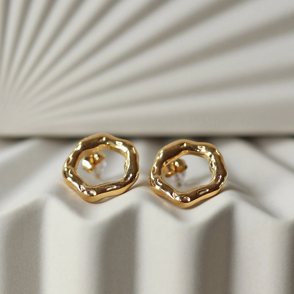 Zoom sur les boucles d'oreilles martelées Source en acier inoxydable doré, posées sur un fond texturé beige.