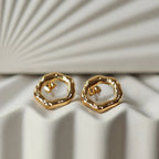 Zoom sur les boucles d'oreilles martelées Source en acier inoxydable doré, posées sur un fond texturé beige.