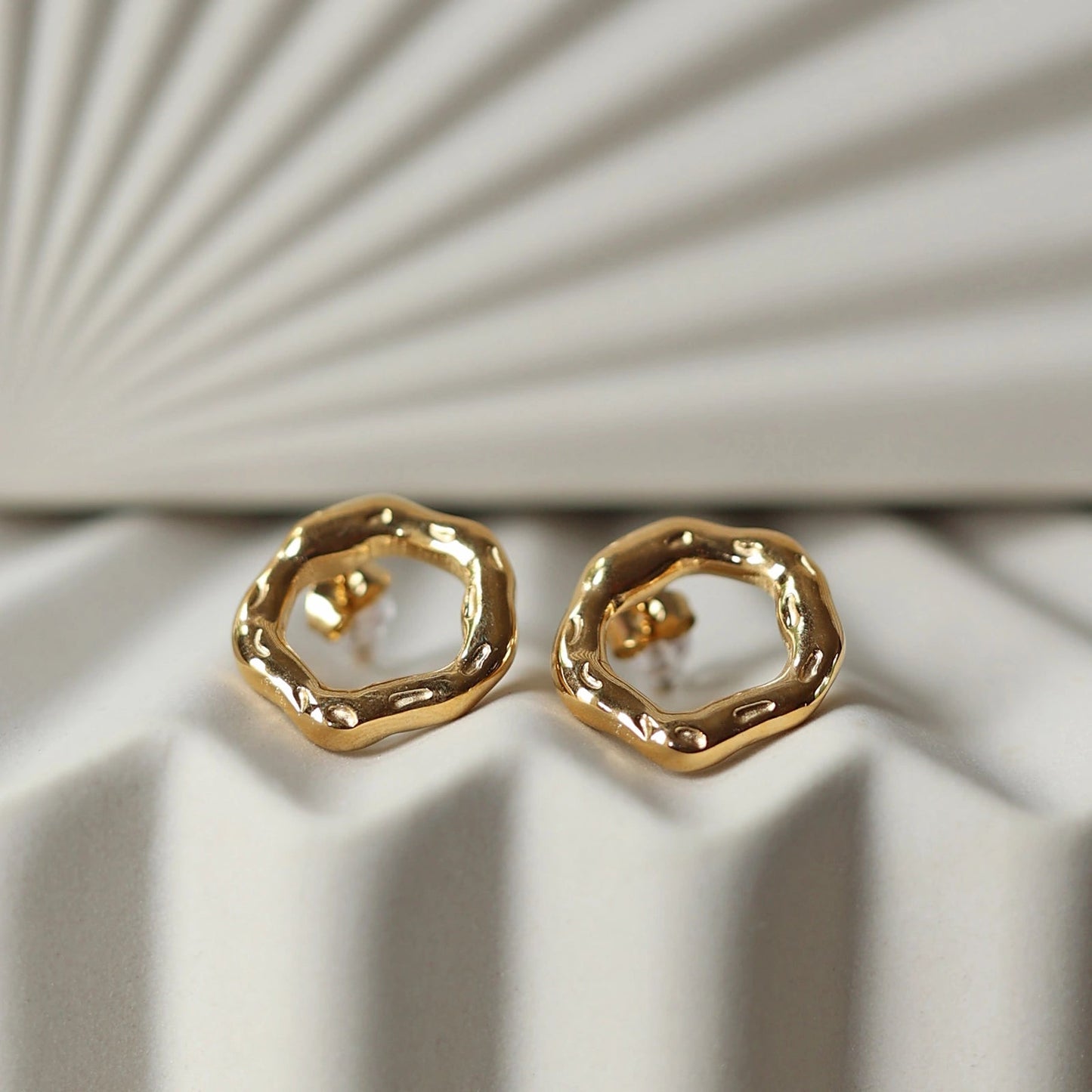 Zoom sur les boucles d'oreilles martelées Source en acier inoxydable doré, posées sur un fond texturé beige.