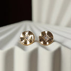 Vue rapprochée des boucles d’oreilles Céleste dorées motif étoile, serties d'un zircon, posées sur un fond beige.