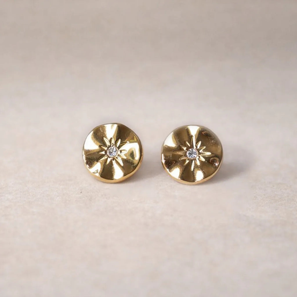 Boucles d’oreilles Minuit dorées motif étoile, serties d'un cristal brillant, posées sur un fond beige - Visée Bijoux.