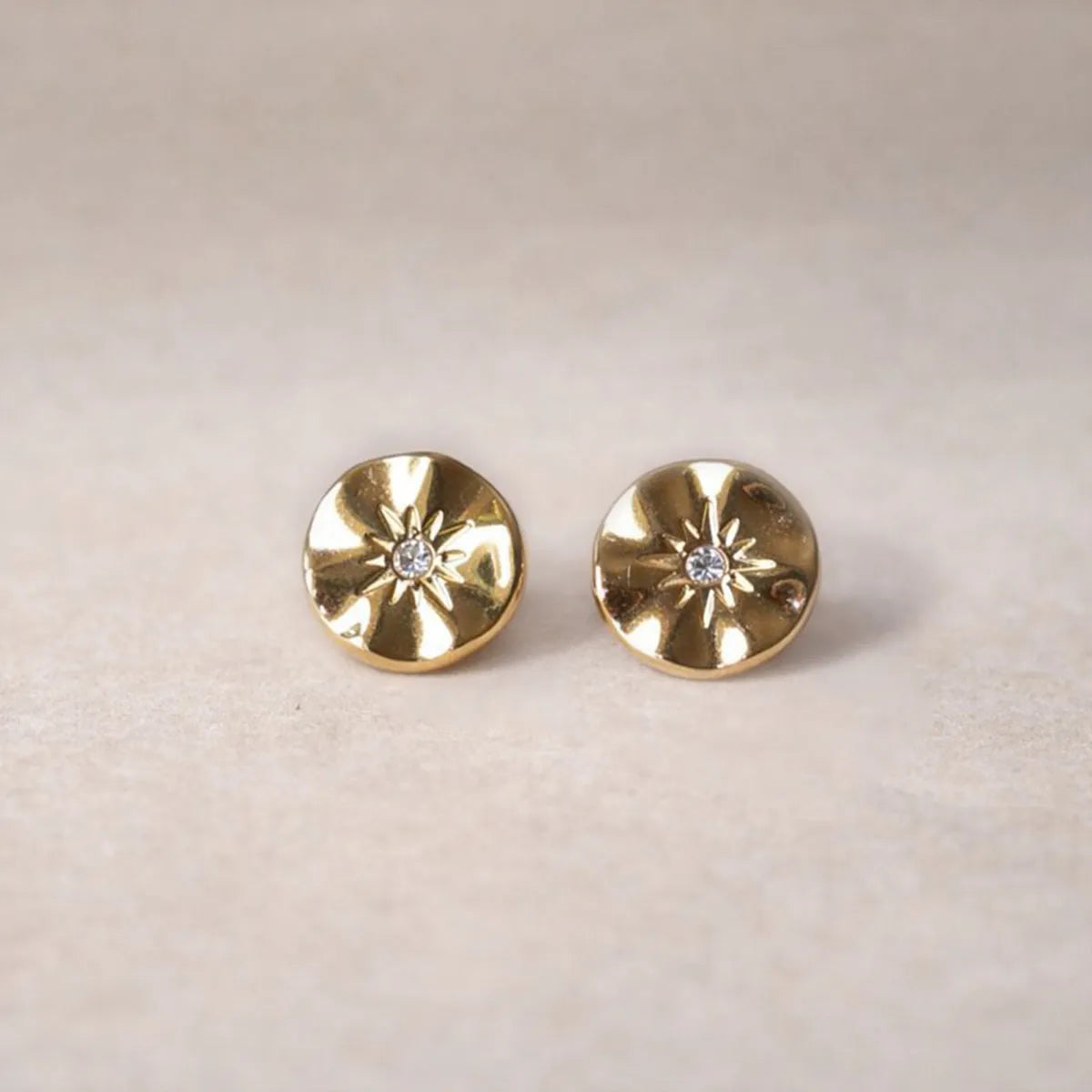 Boucles d’oreilles Minuit dorées motif étoile, serties d'un cristal brillant, posées sur un fond beige - Visée Bijoux.