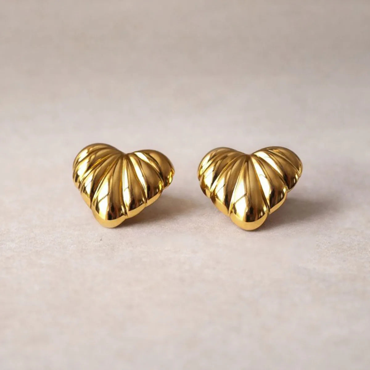 Boucles d'oreilles "Coeur audacieux", en acier inoxydable doré, au design en relief en forme de coeur, posées côte à côte sur un fond beige texturé.