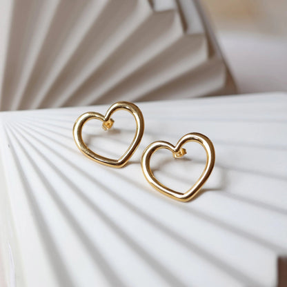 Boucles d'oreilles Douce force dorées, grand cœur en acier, posées sur un support beige texturé.
