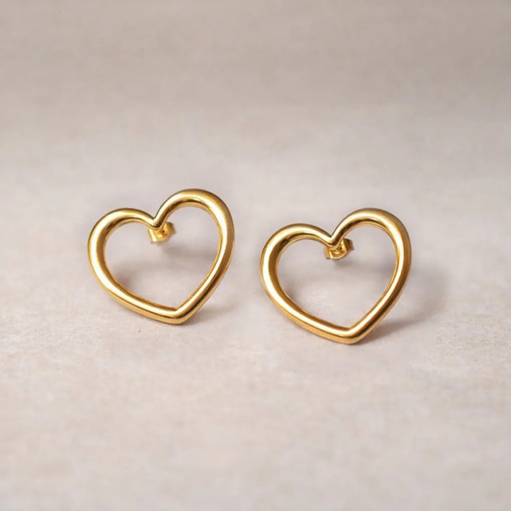 Boucles d'oreilles Douce force dorées, grand cœur en acier, posées sur un fond beige texturé.