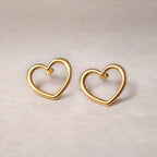 Boucles d'oreilles Douce force dorées, grand cœur en acier, posées sur un fond beige texturé.