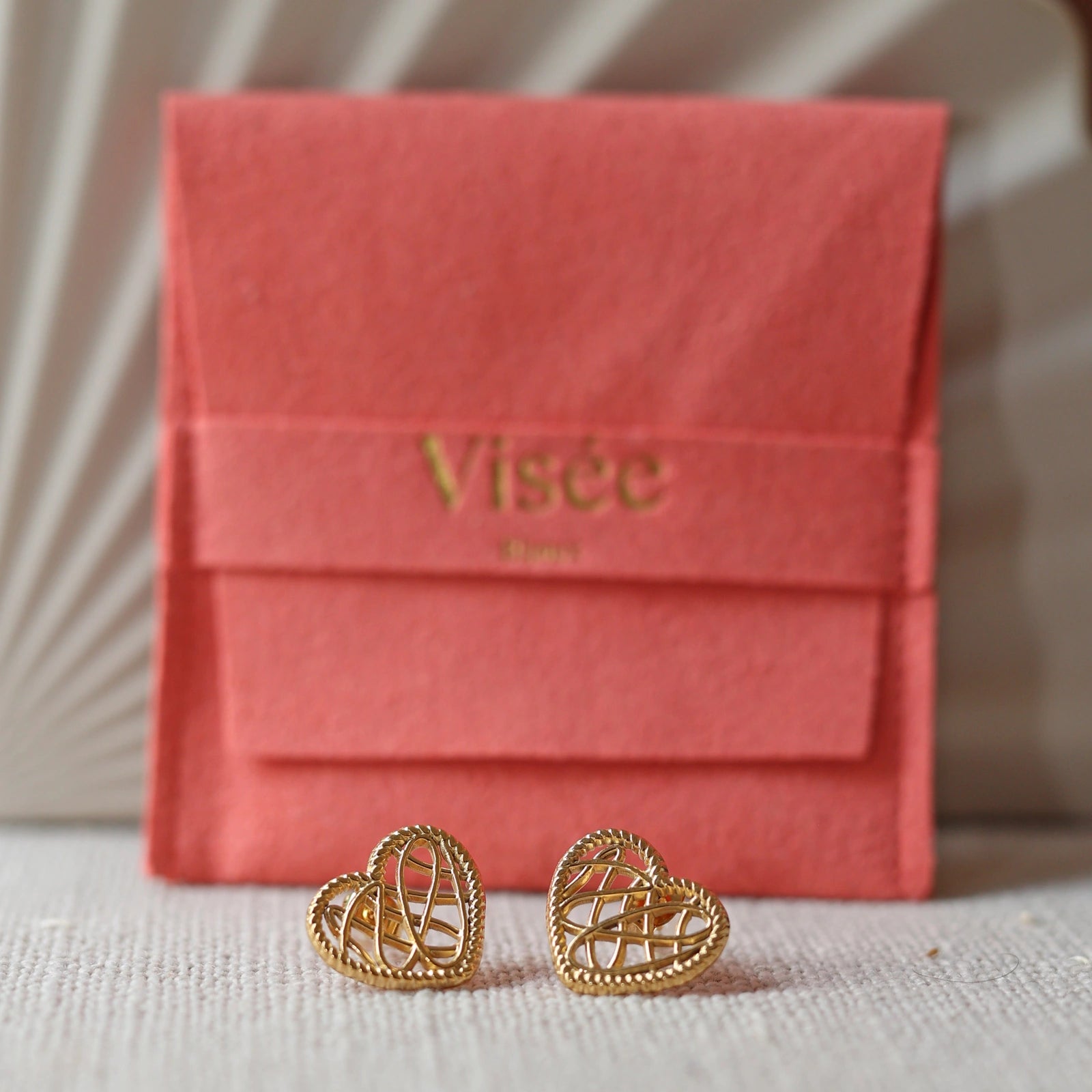 Boucles d'oreilles Etreinte en acier doré, cœurs tressés, posées sur un tissu beige texturé devant le pochon Visée Bijoux. 