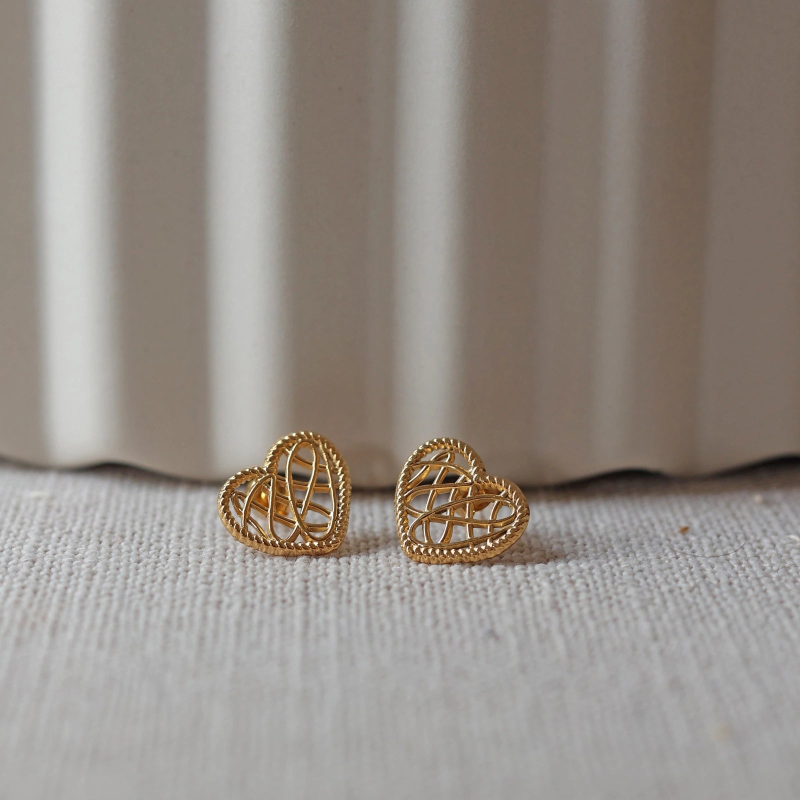 Boucles d'oreilles Etreinte en acier doré, cœurs tressés, posées sur un tissu beige texturé.