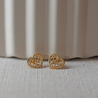 Boucles d'oreilles Etreinte en acier doré, cœurs tressés, posées sur un tissu beige texturé.