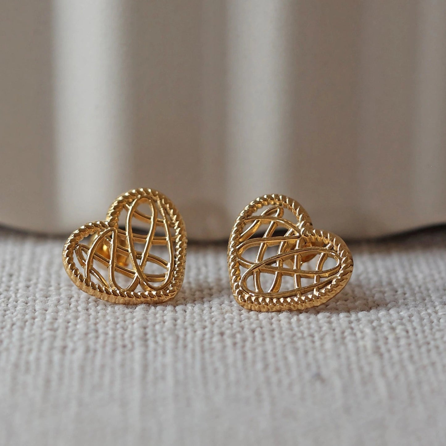 Boucles d'oreilles Etreinte en acier doré, cœurs tressés capturés en gros plan sur un tissu beige texturé.