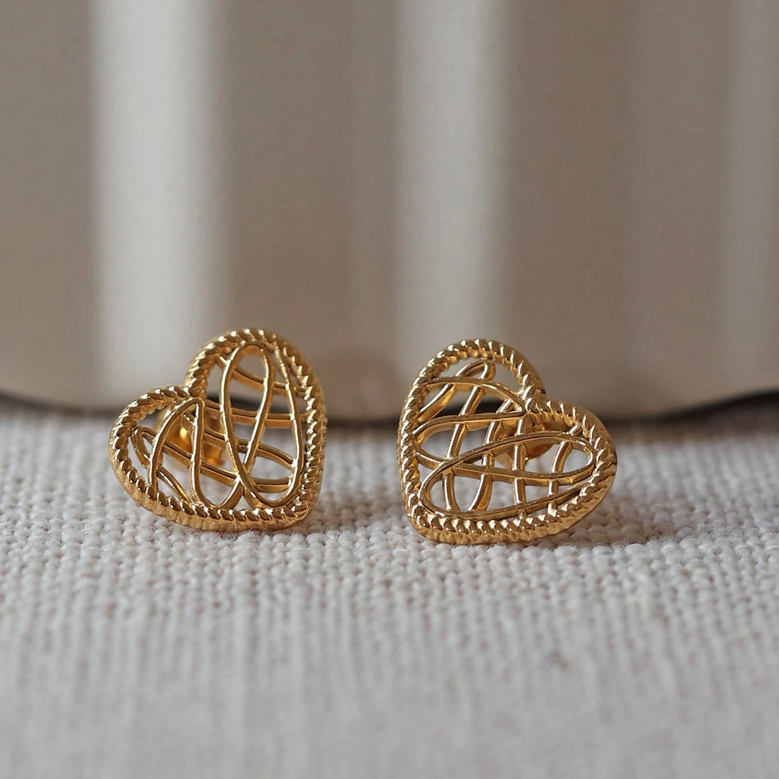 Boucles d'oreilles Etreinte en acier doré, cœurs tressés capturés en gros plan sur un tissu beige texturé.