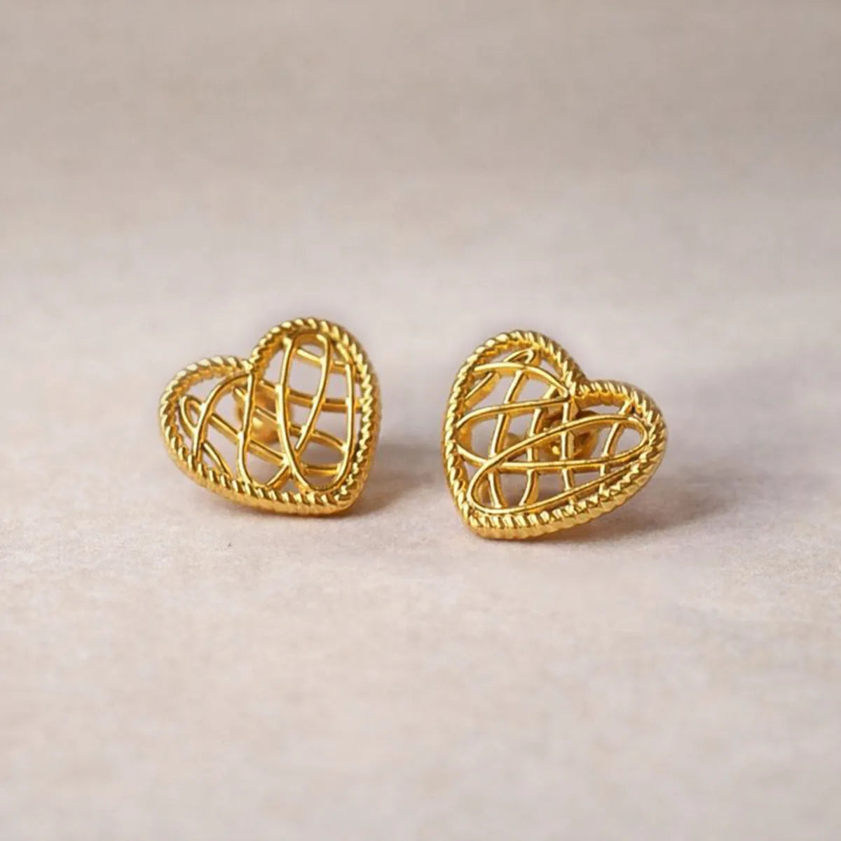 Boucles d'oreilles Etreinte en acier doré, cœurs tressés photographiées en gros plan sur un tissu beige texturé.