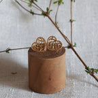 Boucles d'oreilles Etreinte en acier doré, cœurs tressés capturés en gros plan sur un morceau de bois et sur fond de tissu beige texturé et une branche de feuillage