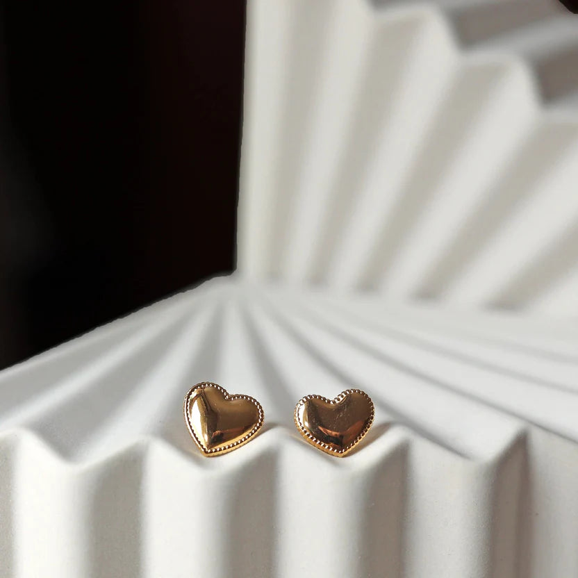 Boucles d'oreilles "L'intime" en forme de cœur doré à contour dentelé, en acier inoxydable, posées sur un fond beige texturé.