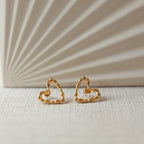 Boucles d'oreilles Insolente Douceur, en forme de cœur martelé, en acier inoxydable doré, photographiées à plat sur un tissu beige texturé.