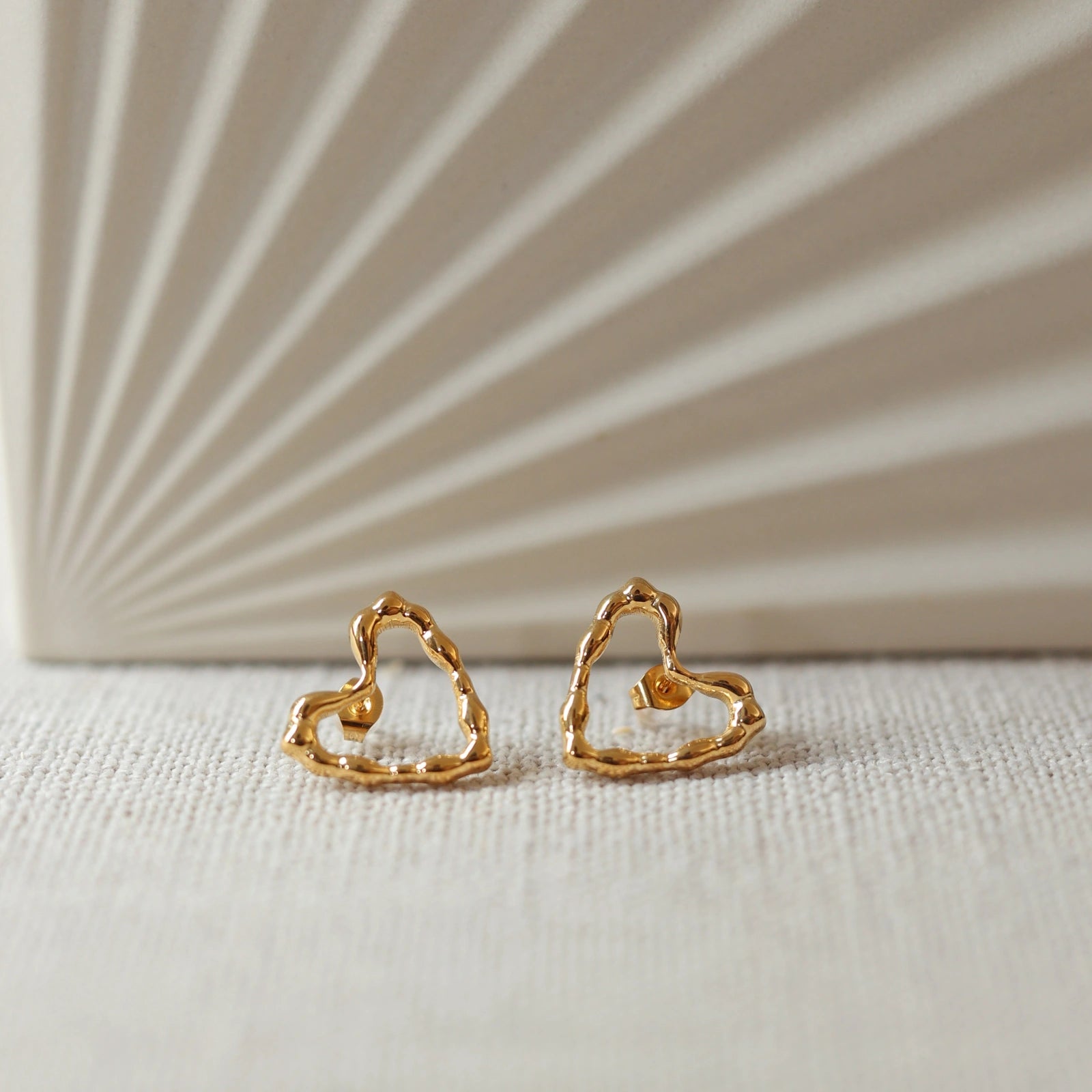 Boucles d'oreilles Insolente Douceur, en forme de cœur martelé, en acier inoxydable doré, photographiées à plat sur un tissu beige texturé.