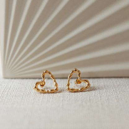Boucles d'oreilles Insolente Douceur, en forme de cœur martelé, en acier inoxydable doré, photographiées à plat sur un tissu beige texturé.
