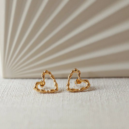 Boucles d'oreilles Insolente Douceur, en forme de cœur martelé, en acier inoxydable doré, photographiées à plat sur un tissu beige texturé.