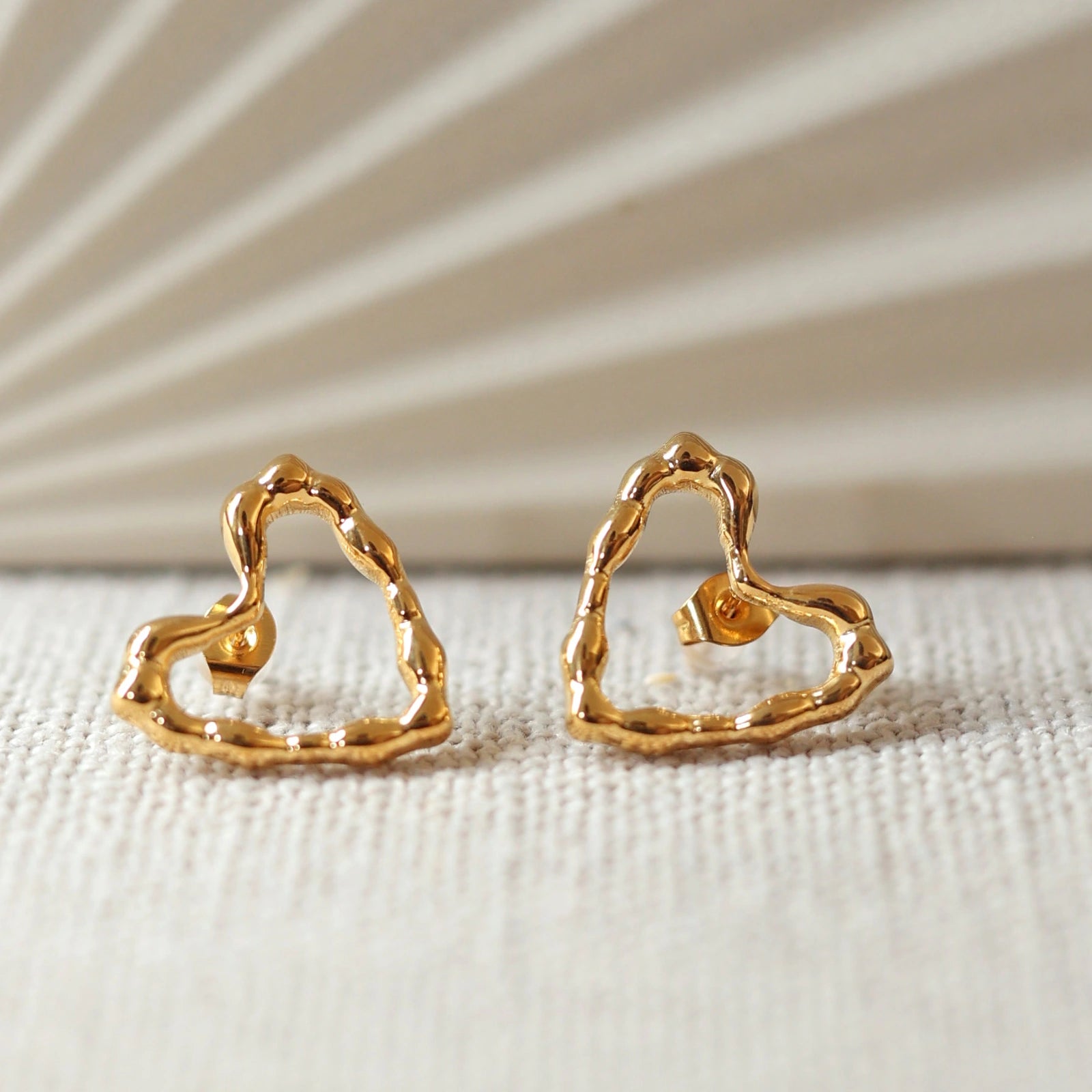 Boucles d'oreilles Insolente Douceur par Visée Bijoux, en forme de cœur martelé, en acier inoxydable doré, vue en gros plan sur un tissu beige texturé.