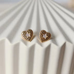 Boucles d'oreilles Promesse, en forme de cœur ornées d'une étoile, en acier inoxydable doré, posées sur un support beige texturé.