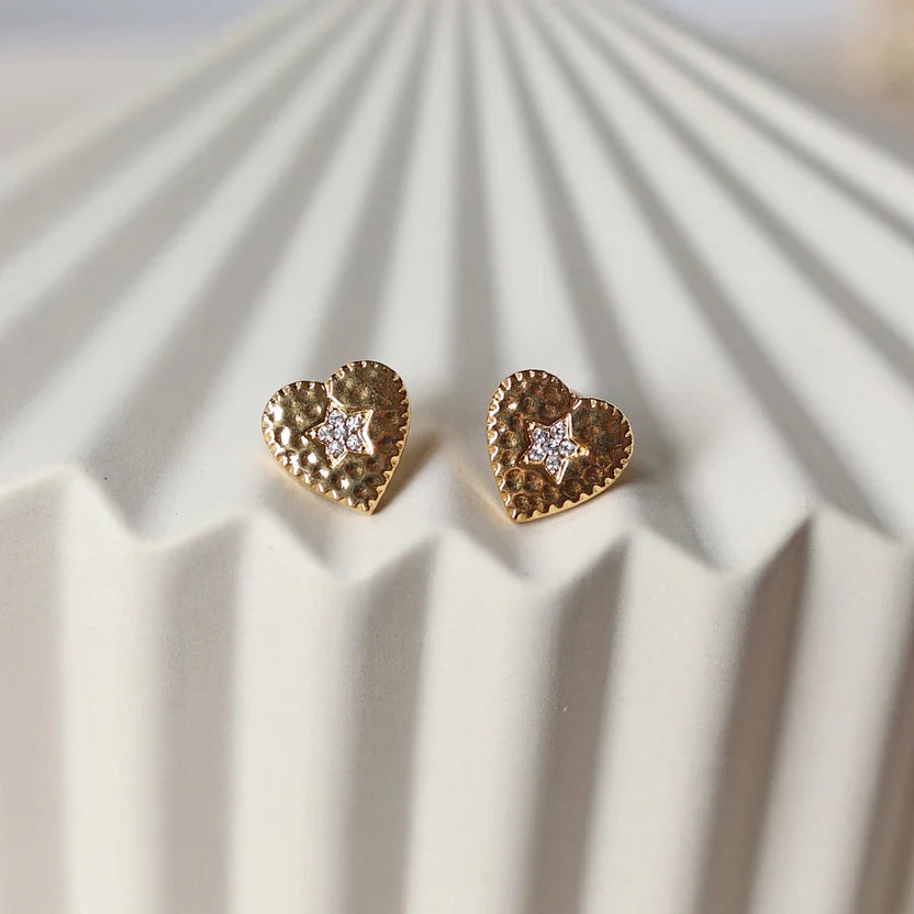 Boucles d'oreilles Promesse, en forme de cœur ornées d'une étoile, en acier inoxydable doré, posées sur un support beige texturé.