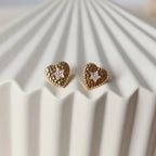 Boucles d'oreilles Promesse, en forme de cœur ornées d'une étoile, en acier inoxydable doré, posées sur un support beige texturé.
