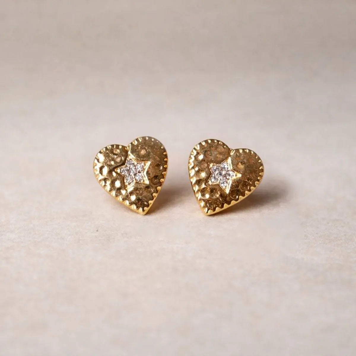 Boucles d'oreilles Promesse, en forme de cœur ornées d'une étoile, en acier inoxydable doré, posées sur un fond beige texturé.