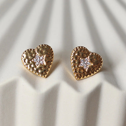 Boucles d'oreilles Promesse, en forme de cœur ornées d'une étoile, en acier inoxydable doré, photographiées en gros plan sur un support beige texturé.
