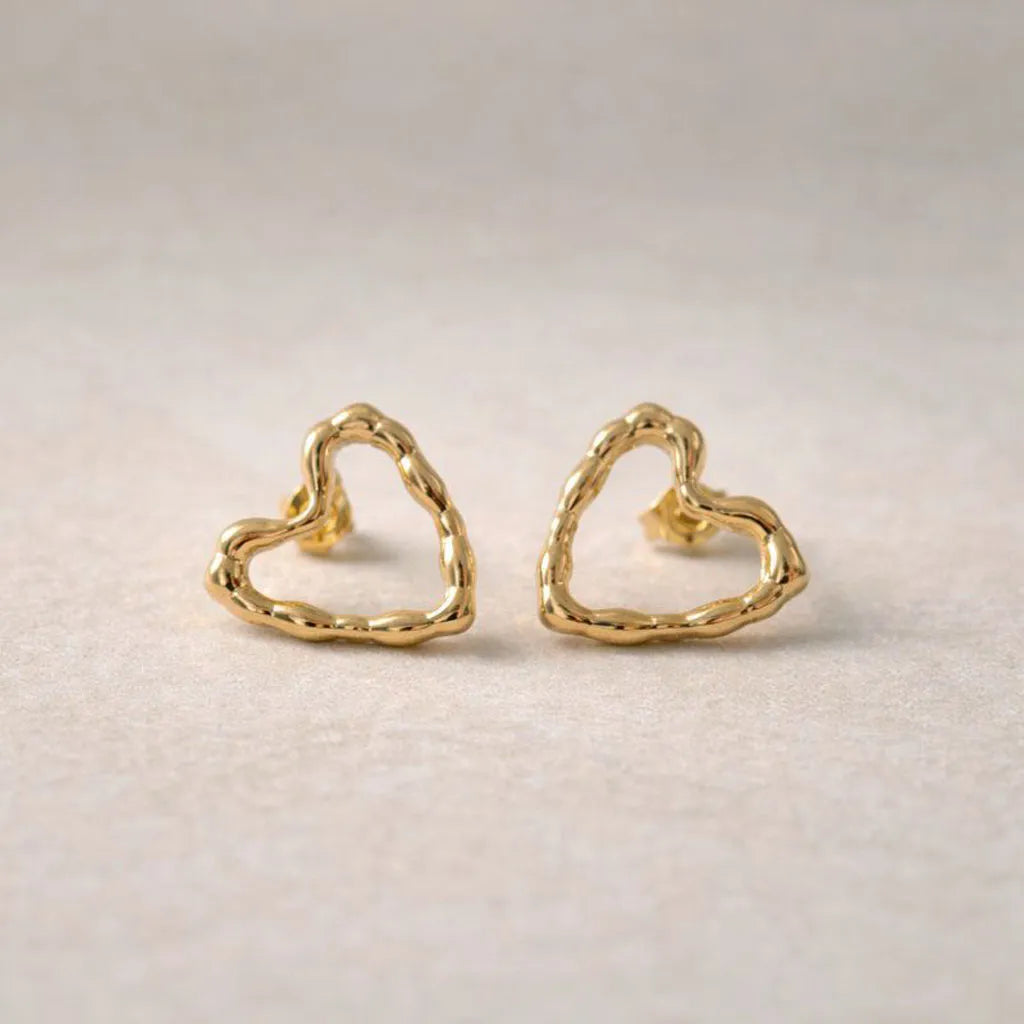 Boucles d'oreilles charismatique, en forme de cœur martelé, en acier inoxydable doré, photographiées à plat sur un tissu beige texturé.