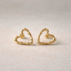 Boucles d'oreilles charismatique, en forme de cœur martelé, en acier inoxydable doré, photographiées à plat sur un tissu beige texturé.