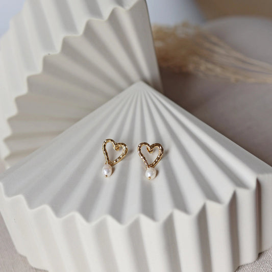 Boucles d’oreilles Loyale, en forme de cœur doré avec perle blanche, en acier inoxydable, photographiée en vue d'ensemble sur un fond beige texturé. 