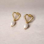 Boucles d’oreilles Loyale, en forme de cœur doré avec perle blanche, en acier inoxydable, photographiée en vue d'ensemble sur un fond beige texturé. 