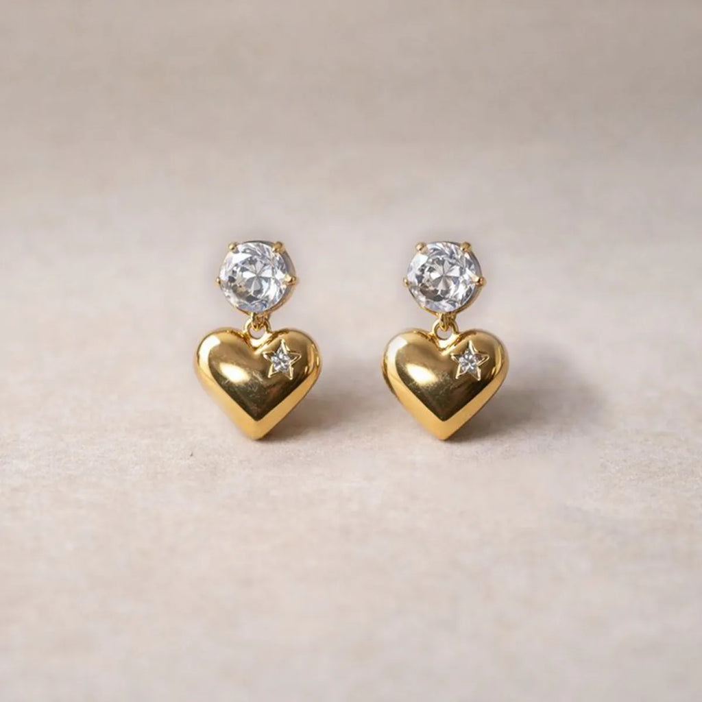 Boucles d'oreilles Intuition en acier doré, cœur délicat serti de pierre scintillante, posées sur un fond beige texturé