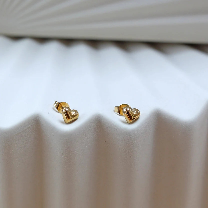 Zoom sur les boucles d'oreilles "Tendre discrétion" de Visée, en forme de cœur en acier doré, posées sur un fond beige texturé.