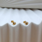 Zoom sur les boucles d'oreilles "Tendre discrétion" de Visée, en forme de cœur en acier doré, posées sur un fond beige texturé.