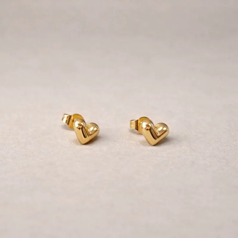 Zoom sur les boucles d'oreilles "Tendre discrétion" de Visée, en forme de cœur en acier doré, posées sur un fond beige texturé.