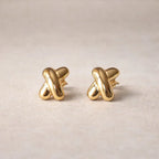 Boucles d'oreilles Nouée, au design croix en acier inoxydable doré, photographiées sur un fond beige texturé.