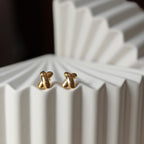 Boucles d'oreilles "Révèle toi", au design croix en acier inoxydable doré, photographiées sur un fond beige texturé.