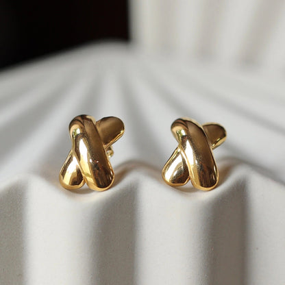 Boucles d'oreilles "Révèle toi", au design croix en acier inoxydable doré, photographiées sur un fond beige texturé.